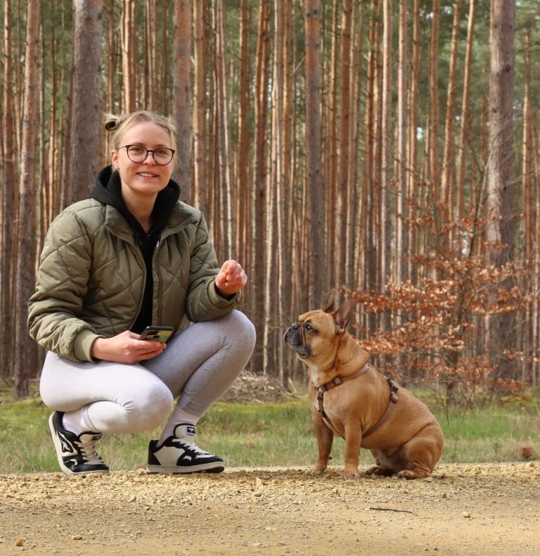 Frau kniet im Wald neben einem Hund und hält ein Leckerli in der Hand.